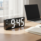 Digital alarm clock on a bright work desk next to a notebook and laptop during the day