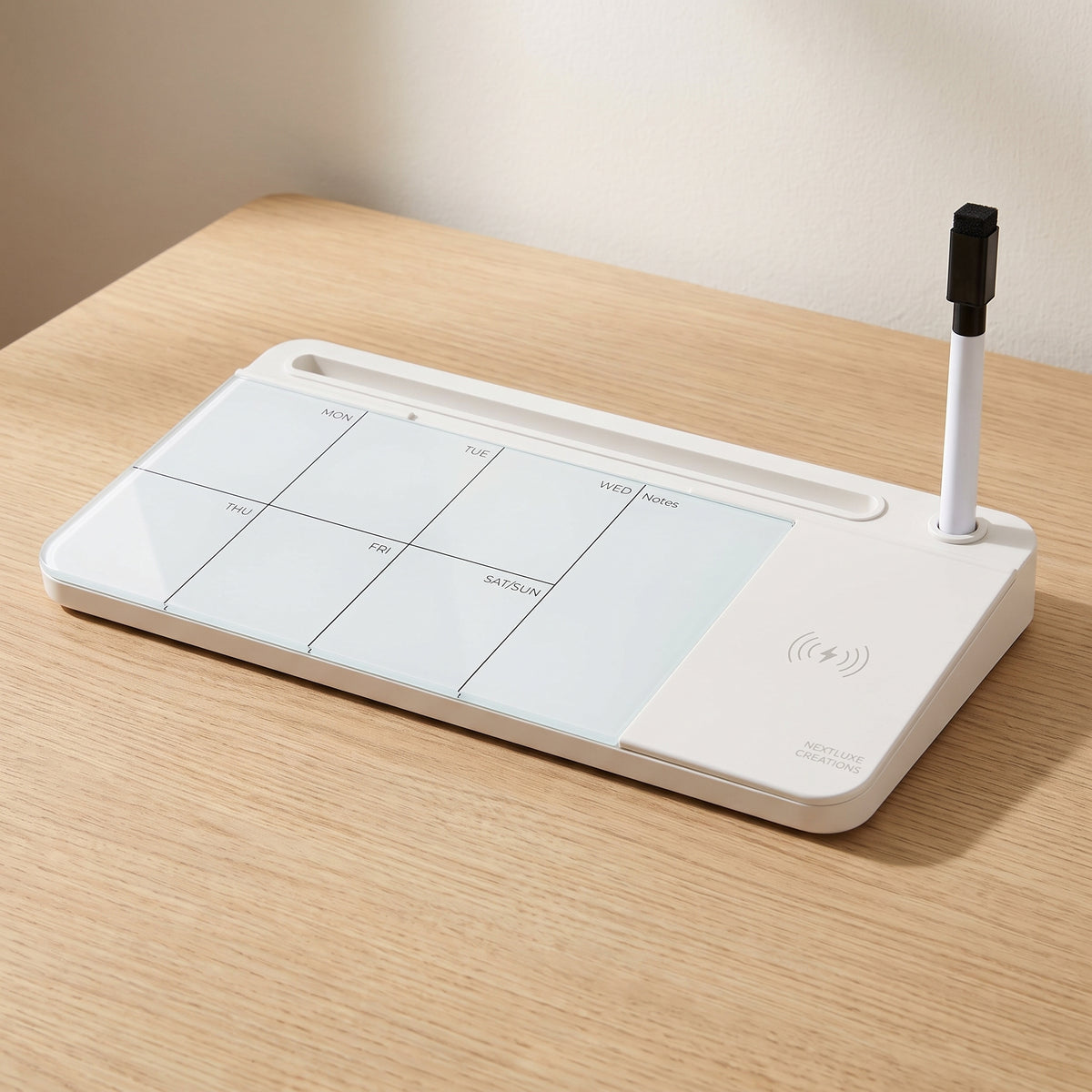 White glass desk whiteboard on a pale oak desk, its weekly grid on the left and the wireless charging pad with its glyph on the right